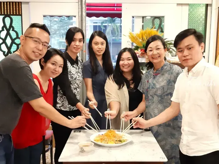 Group of seven people joyfully tossing yusheng with chopsticks during a festive celebration at a restaurant.