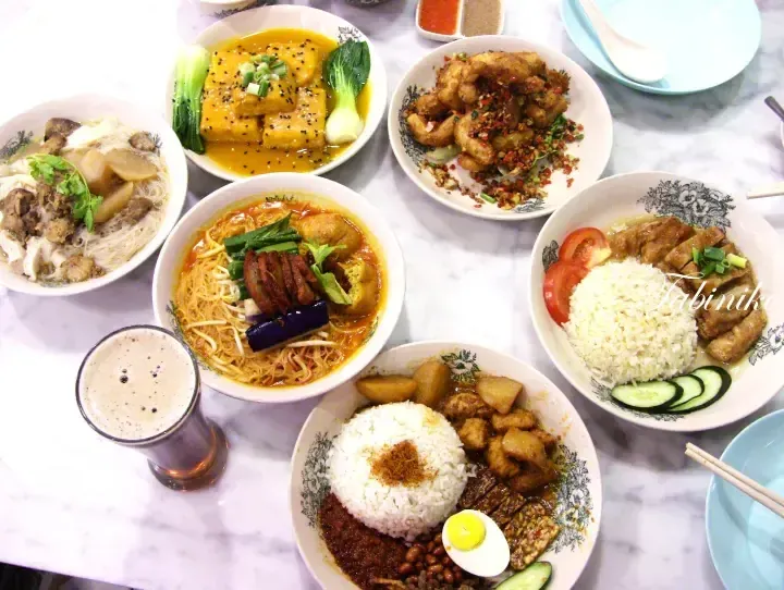 A table spread featuring Malaysian dishes like nasi lemak, laksa, and curry tofu alongside a drink.