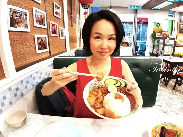 Woman in red top holding a plate of Nasi Lemak with chicken and cucumber at a restaurant.