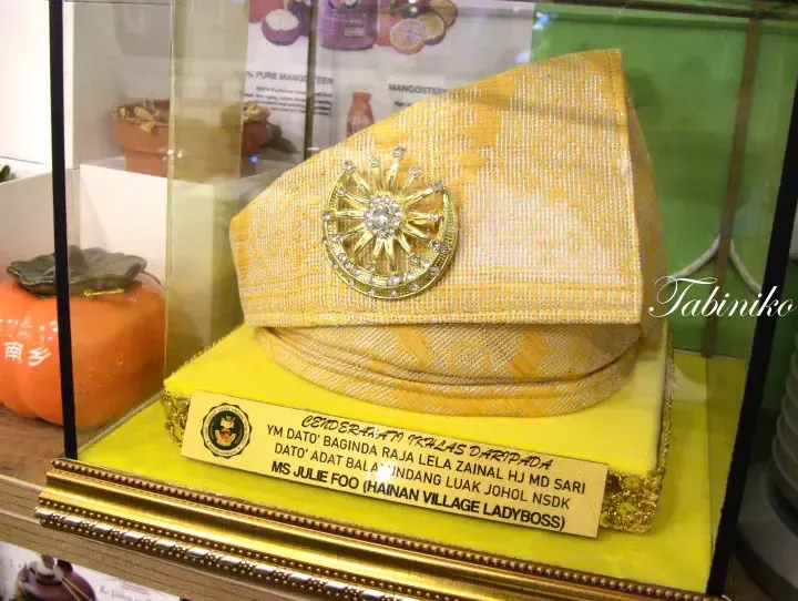 Yellow traditional Malay songkok with a gold emblem displayed in a glass case with a gift plaque.