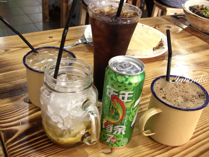 マレーシアの食堂で木製テーブルに並ぶ7up缶やエナメルマグのカフェなど様々なアイスドリンク