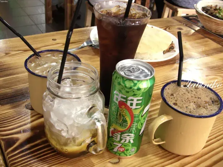 Assorted iced drinks including a 7up can and coffee served in enamel mugs on a wooden table at a Malaysian eatery