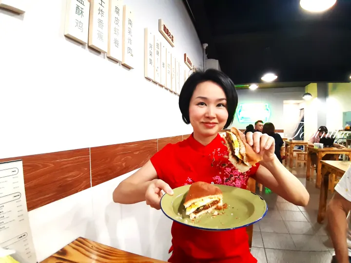 壁にメニューボードがあるマレーシアのカフェで、赤いチャイナドレスを着た女性がサンドイッチを持っている。