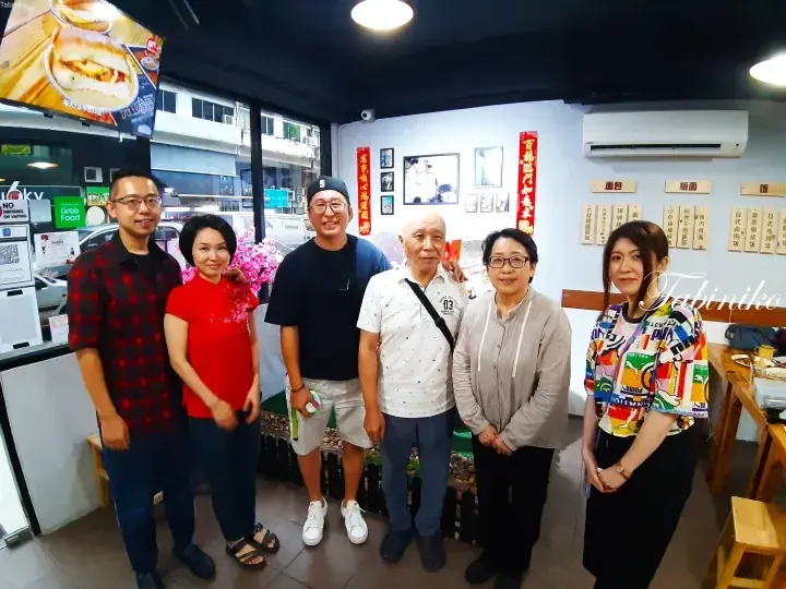 A group of six people posing for a photo inside a restaurant with menu boards and Grab Food signage visible in the background.