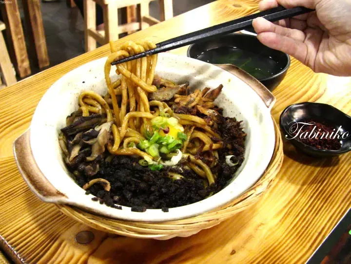 Thick noodles topped with minced meat, mushrooms, and green onions being lifted with chopsticks in a ceramic bowl.