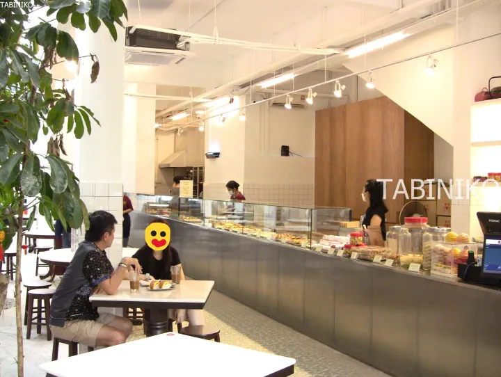 パントリーやパンが並ぶガラスケースを前に、明るいカフェで食事をする客の姿。