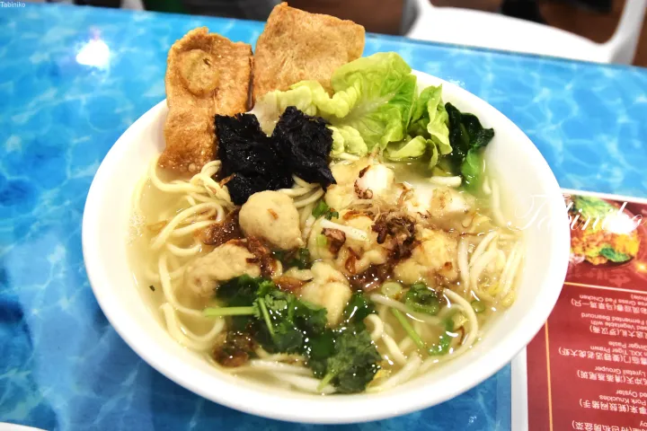 青いテーブルの上に置かれた、油揚げやレタス、ハーブを添えた魚肉団子麺のボウル。