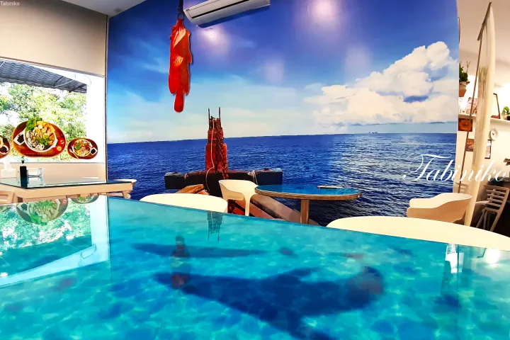 海を模した壁紙と青いテーブルが特徴的なレストランのインテリア。赤い魚のオブジェが吊るされている。