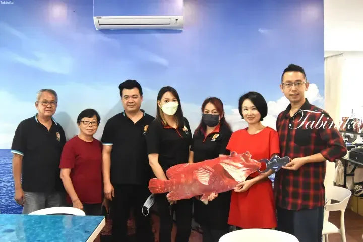 Group of seven people posing with a large red grouper fish cutout inside a restaurant with ocean-themed wall decor.