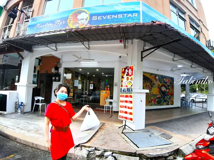赤いドレスを着た女性が「Restoran Master Ng&rsquo;s Sevenstar」の前でテイクアウトバッグを持っている。