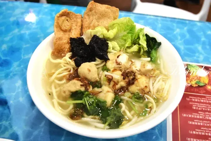 A bowl of fish ball noodles with fried tofu, lettuce, and herbs on a blue table.