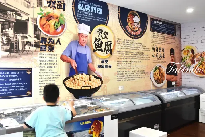 A young boy looks at a display freezer in a shop featuring a large wall mural of Master Ng cooking Golden Yufoo and curry noodles.