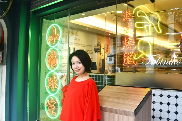 「小龍冰室」と「港式燒臘」のネオンサインが輝くレストランの外でポーズをとる赤い服の女性。