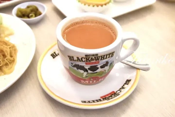 A cup of milky coffee in a Black & White branded mug and saucer, served alongside noodles and a small side dish.