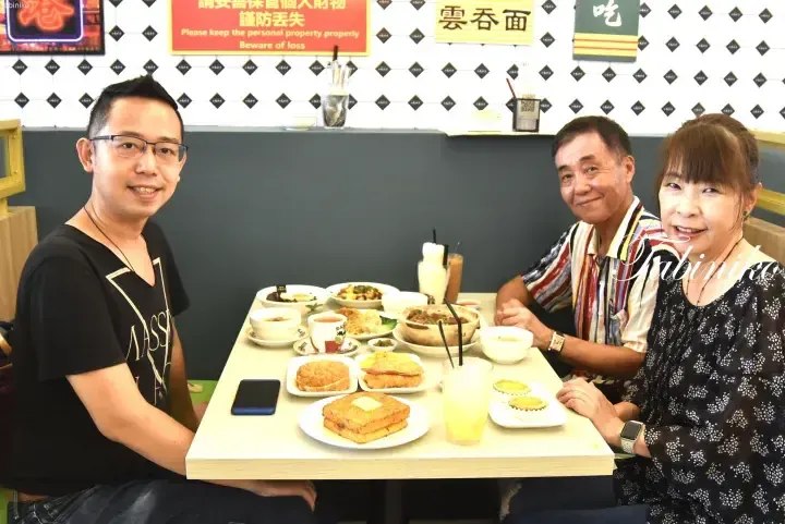 Three people enjoy a meal at a restaurant with dim sum and drinks, featuring a &lsquo;Wonton Noodle&rsquo; sign and a &lsquo;Please keep the personal property properly&rsquo; notice.