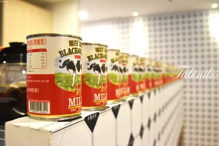 Rows of Black & White condensed milk cans displayed on a shelf with a geometric patterned background.