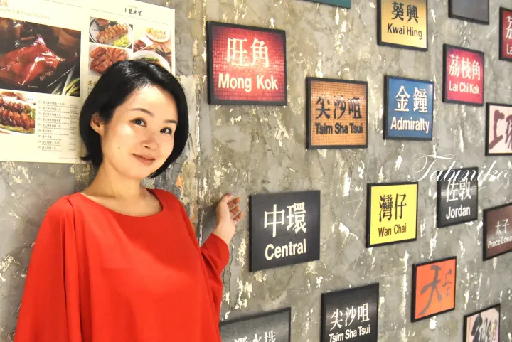 赤いトップスを着た女性が、Mong KokやCentralなどの香港の地名が書かれたカラフルな看板で飾られた壁の前でポーズをとっている。