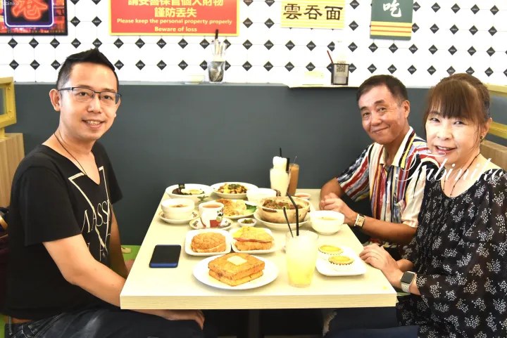 「雲吞面」の看板があるレストランで、点心や飲み物を囲んで食事を楽しむ3人。壁には「Please keep the personal property properly」の注意書きが掲げられている。