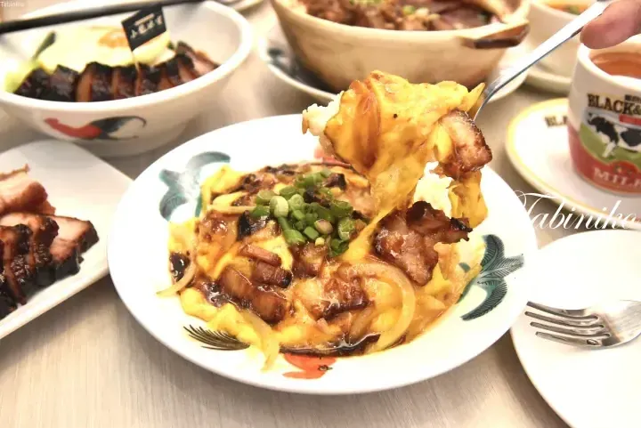 A spoon lifts a bite of fluffy omelette with char siu and onions from a plate, alongside a bowl of rice and a Black & White milk tea.