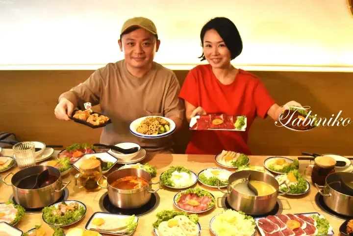 A couple smiles while holding plates of raw meat and fried food at a table filled with hot pot ingredients and broth.