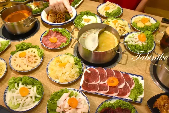 A table filled with various hot pot ingredients including sliced meats, lotus root, and vegetables surrounding a split broth pot.