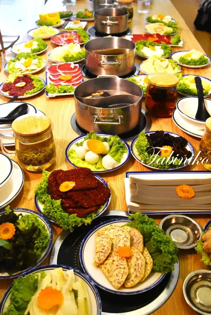 A long wooden table set with multiple hot pot stations and plates of fresh ingredients like meat, vegetables, and noodles.