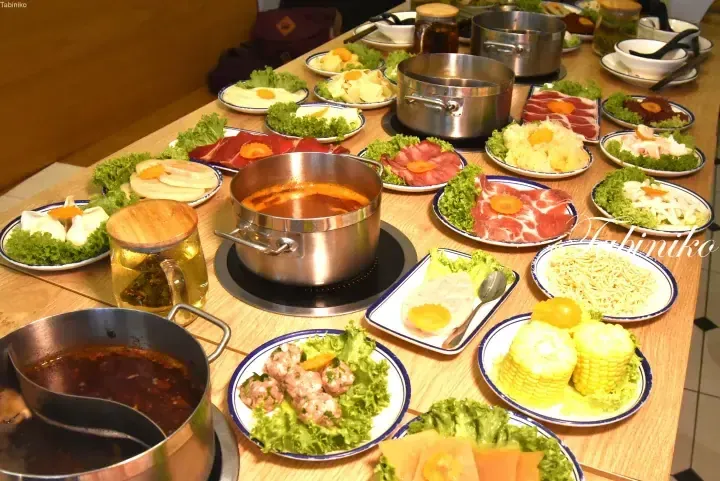 A table filled with various hot pot ingredients including meats, vegetables, corn, and noodles alongside simmering pots.