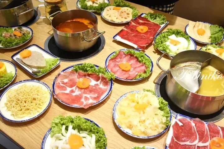 A table spread with various hot pot ingredients including sliced meats, vegetables, noodles, and two simmering pots.