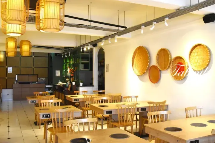 Interior of a modern restaurant featuring wooden tables with built-in hot pot burners, bamboo pendant lights, and woven baskets on the wall.