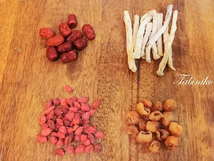 Four piles of traditional Chinese herbal ingredients including red dates, goji berries, dried longan, and astragalus root on a wooden surface.