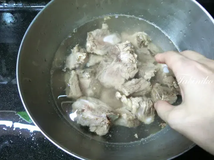 A hand placing pork ribs into a pot of water for boiling, a common step in preparing Malaysian dishes.