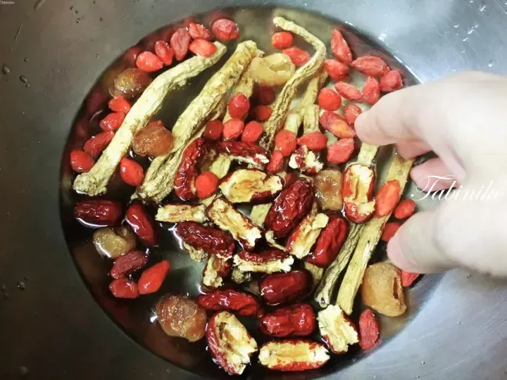 A hand adds ingredients like red dates, goji berries, and herbs into a metal bowl of water for traditional Chinese herbal soup.