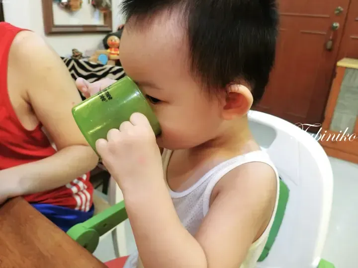 A toddler in a white tank top drinks from a green cup while seated in a high chair at home.