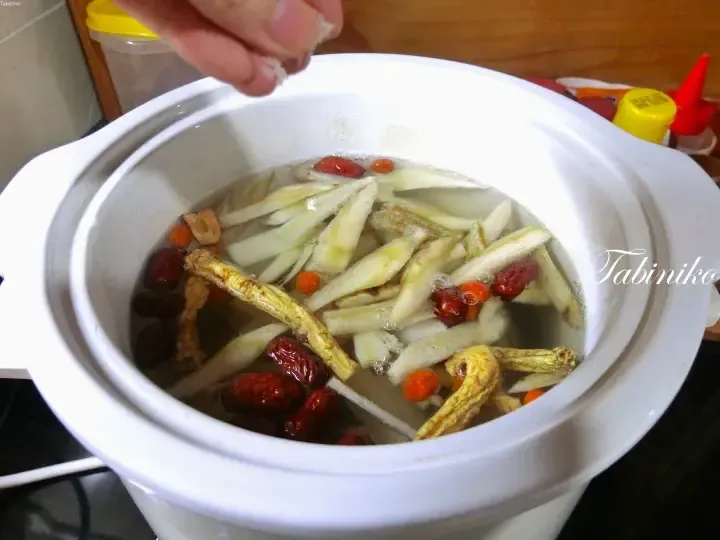 A hand sprinkles seasoning into a white ceramic pot containing herbal soup with ginseng, red dates, and goji berries.