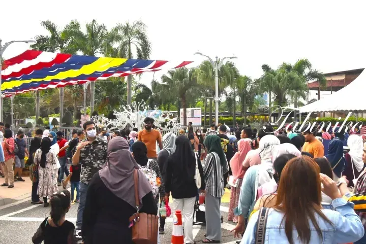 Crowd walking under colorful Malaysian flags at a festive outdoor event with palm trees.