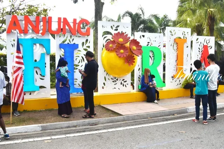 Anjung Floria sign with colorful letters and a yellow sun, with visitors posing near a Malaysian flag.