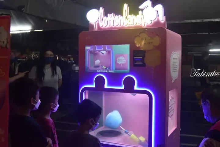 Children gather around a pink Cotton Candy vending machine at a night market, watching the automated process.