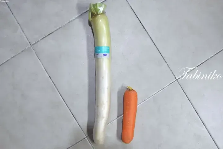 A long white radish with a green label and a carrot placed on a tiled floor.