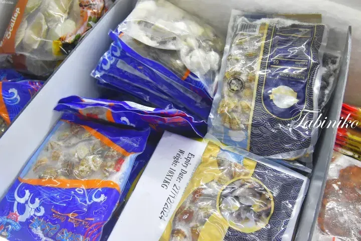 Assorted frozen seafood packages in a cooler box, including clams and scallops with visible expiry date labels.