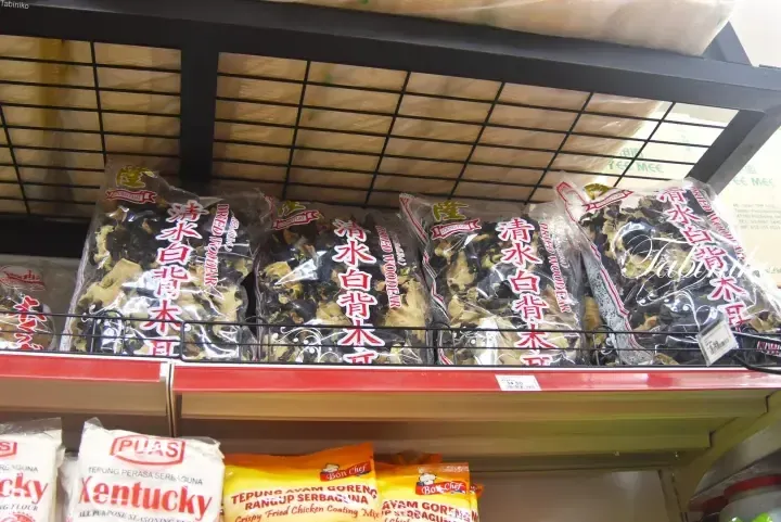 Shelves stocked with Loong Fuat dried wood ear mushrooms and PUAS Kentucky seasoning flour in a Malaysian grocery store.