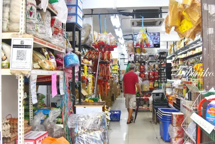 A man walks through a narrow aisle in a Malaysian grocery store filled with hanging snacks and shelves of packaged goods.