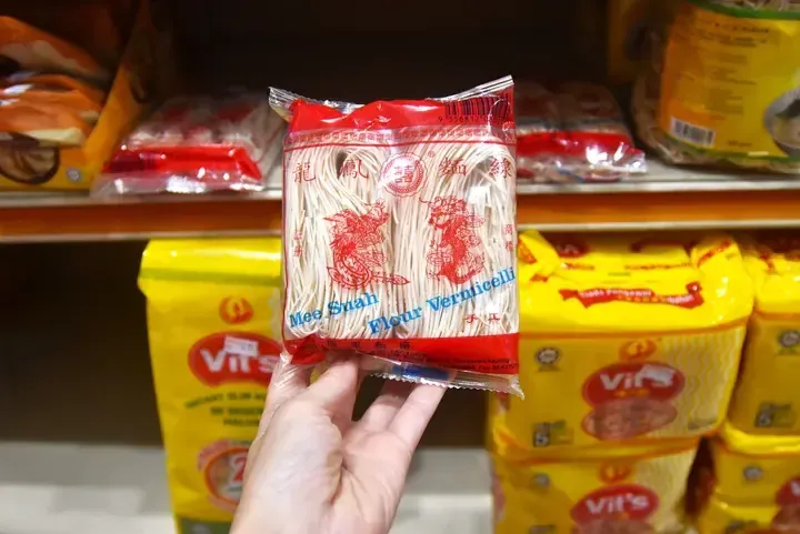 A hand holds a red package of Long Fung Mee Suah Flour Vermicelli noodles on a grocery store shelf.
