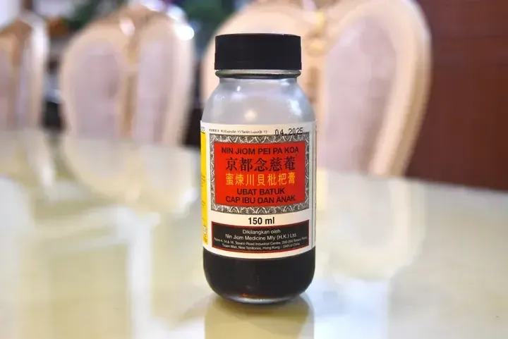 A bottle of Nin Jiom Pei Pa Koa cough syrup sits on a table with decorative shells in the background.