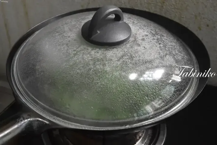 A glass pot lid with condensation covering a pan of cooking green vegetables on a stove.