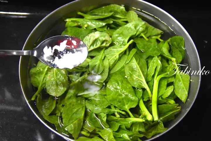 Fresh spinach leaves in a metal bowl with a spoonful of white powder being added.