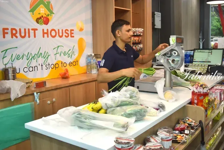 A shop assistant at Fruit House weighs fresh vegetables behind the counter, with the slogan Everyday Fresh You can’t stop to eat visible on the wall.
