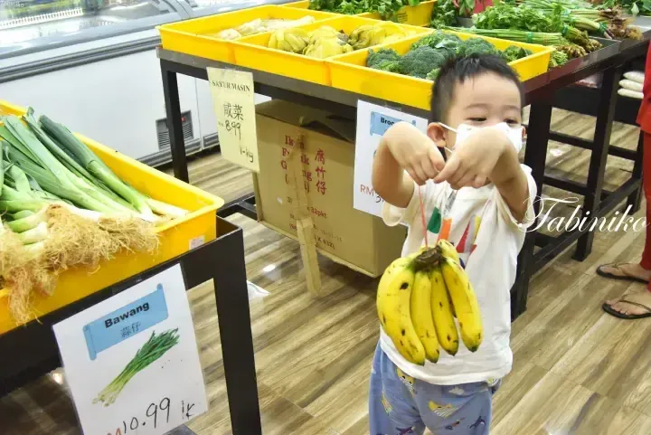 A young boy holds a bunch of bananas in a Malaysian grocery store aisle with yellow vegetable bins and price signs.