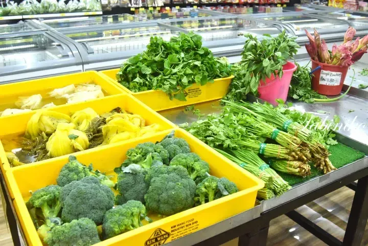 Fresh vegetables including broccoli, pickled mustard greens, and herbs displayed in yellow bins at a Malaysian supermarket.