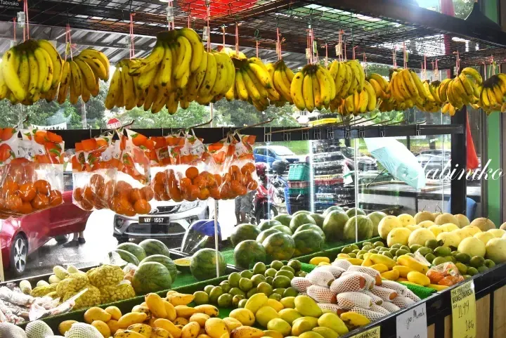 Fresh bananas, oranges, and melons displayed at a fruit stall in Malaysia with cars visible outside.