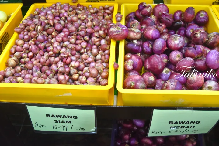マレーシアの市場で販売されている黄色い箱に入ったシアンワタと赤タマネギ、価格札が見える。